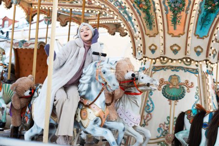 Photo for Cheerful young woman in a faux fur coat and mittens rides a carousel and has fun - Royalty Free Image
