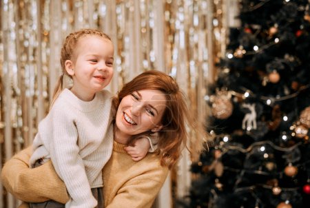 Photo for A young mother holds a laughing daughter in her arms next to the Christmas tree. Family, winter holidays and people concept. - Royalty Free Image