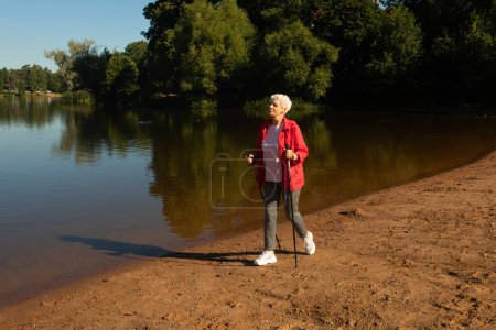 Téléchargez les photos : Femme âgée aux cheveux gris marchant avec des bâtons de suivi sur la plage près du lac - en image libre de droit
