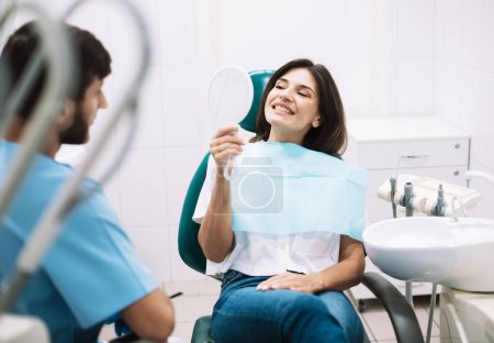 Foto de Mujer joven sosteniendo el espejo y mirando su hermosa sonrisa sentada en el consultorio dental. Feliz sonrisa.. - Imagen libre de derechos