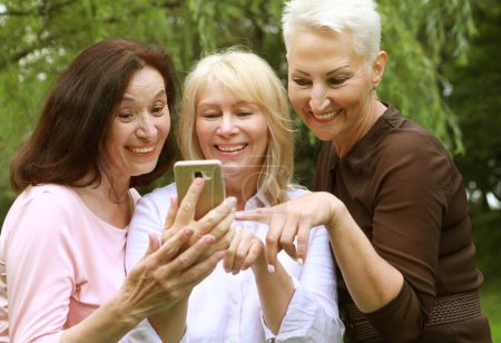 Foto de Three elderly women are smiling and looking photos at the at screen of the phones in the park on a summer day. Golden age. - Imagen libre de derechos