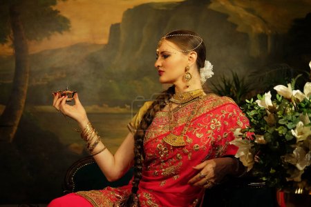 Foto de Charming Indian bride dressed in Hindu red traditional wedding clothes embroidered with gold sitting on sofa near flowers, smiles and holding small gold box. - Imagen libre de derechos