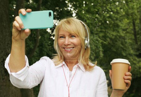 Foto de Charming senior blond woman holding a cup of coffee to go and making selfie in summer park. Lifestyle and gold age concept. - Imagen libre de derechos