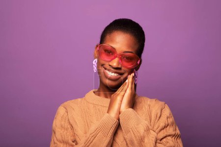 Téléchargez les photos : Charmante jeune femme noire à la mode des années 20 porte un pull beige et de grandes lunettes de soleil sur fond violet - en image libre de droit