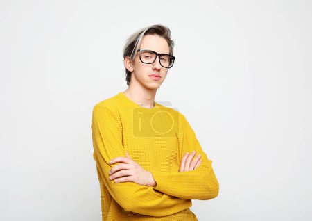 Foto de Un joven con un suéter amarillo y gafas posa con los brazos cruzados sobre un fondo gris. Primer plano retrato. - Imagen libre de derechos