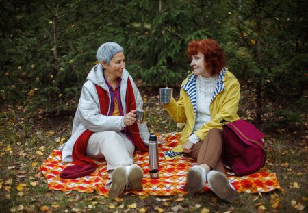 Téléchargez les photos : Deux femmes âgées sont assises sur une couverture à carreaux dans la forêt, buvant du café, parlant, s'amusant. - en image libre de droit