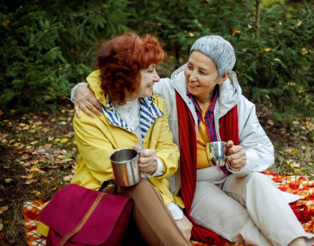 Foto de Dos ancianas están sentadas en una manta a cuadros en el bosque, bebiendo café, hablando, divirtiéndose. - Imagen libre de derechos