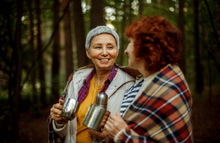 Téléchargez les photos : Deux amies âgées marchent dans la forêt, versent le café d'un thermos, passent un bon moment ensemble. Style de vie et concept de personnes. - en image libre de droit