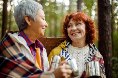 Téléchargez les photos : Deux amies âgées marchent dans la forêt, versent le café d'un thermos, passent un bon moment ensemble. Style de vie et concept de personnes. - en image libre de droit