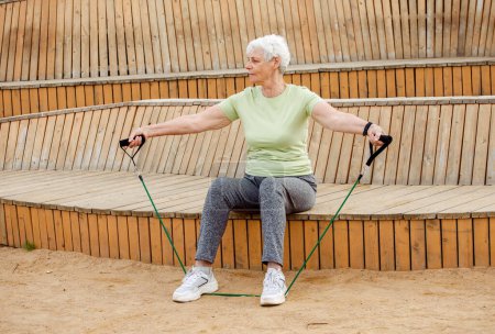 Téléchargez les photos : Vieille femme aux cheveux courts faisant des exercices en plein air, assise sur un banc en bois en utilisant des élastiques de résistance. Concept de style de vie. - en image libre de droit