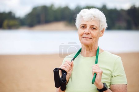 Photo for Smiling old woman wear sportswear doing exercises outdoor on the beach in the morning using resistance rubber bands. Healthy lifestyle, active retired life and sporty time. - Royalty Free Image