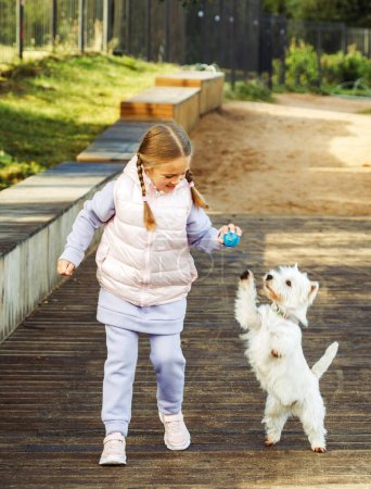 Photo for Seven-year-old blond girl playing with her white puppy in the autumn park, lifestyle concept. - Royalty Free Image