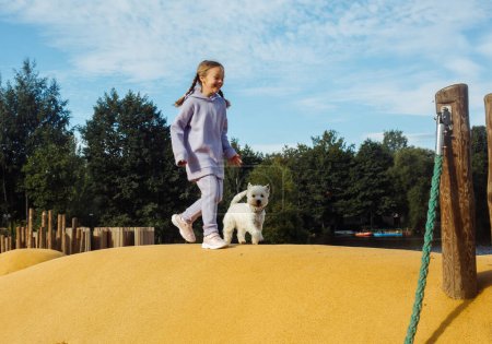 Téléchargez les photos : Fille blonde de sept ans jouant dans l'aire de jeux avec son chiot blanc, jour d'été ensoleillé - en image libre de droit