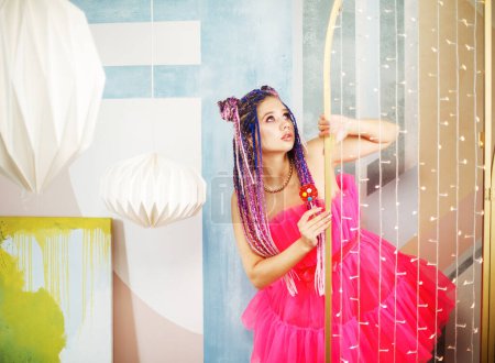 Foto de Joven bonita mujer con rastas peinado usando vestido rosa, estilo muñeca, maquillaje brillante, hasta en la habitación rosa. - Imagen libre de derechos