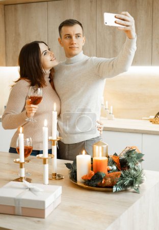 Téléchargez les photos : Jeune couple heureux célébrant Noël, tenant des verres de vin, embrassant, prenant selfie. Style de vie et concept de personnes. - en image libre de droit