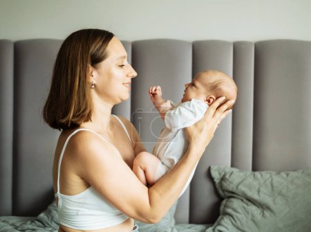Foto de Una joven madre está sentada en la cama y sosteniendo a un bebé en sus brazos. Concepto de estilo de vida y personas. - Imagen libre de derechos