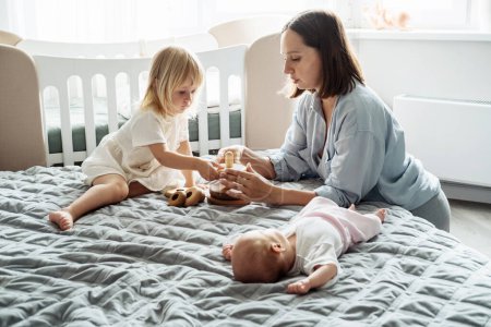 Téléchargez les photos : Une jeune mère avec un bébé sur le lit, à côté d'une fille de deux ans, sœur. Amour et tendresse. - en image libre de droit