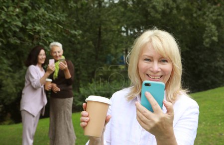 Foto de Beautiful old blond woman laughing, holding a cup of coffee to go, take selfie and feeling so happy. Lifestyle and gold age concept. - Imagen libre de derechos