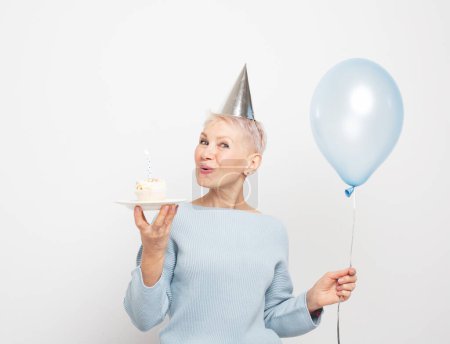 Photo for People, party, aging and maturity concept. Beauitful elderly female wearing conical hat celebrating birthday and blows out the candle on cupcake - Royalty Free Image
