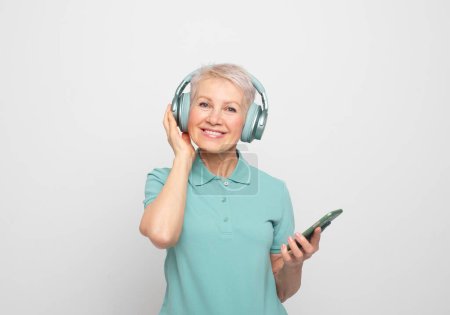 Téléchargez les photos : Charming pensioner wearing wireless headphones. A modern elderly woman listens to music using a smartphone. - en image libre de droit