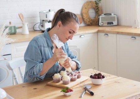 Téléchargez les photos : Cooking, Freelance, People and Hobbies concept: A young woman wearing casual decorates muffins with white cream. - en image libre de droit