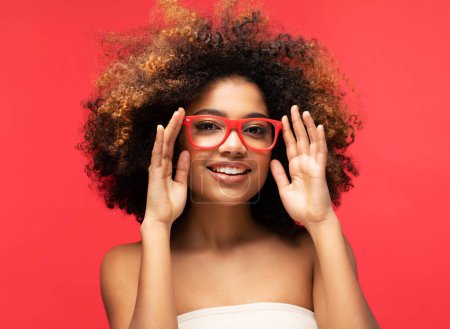 Téléchargez les photos : Heureuse femme afro portant des lunettes souriantes, a une bonne humeur isolée sur fond rouge studio. - en image libre de droit