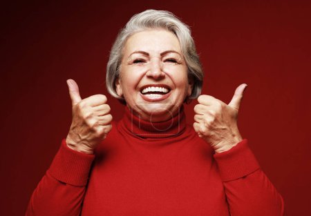 Foto de Estilo de vida, emoción y concepto de la gente: anciana feliz mujer dando un pulgar hacia arriba y mirando a la cámara sobre el fondo rojo. - Imagen libre de derechos