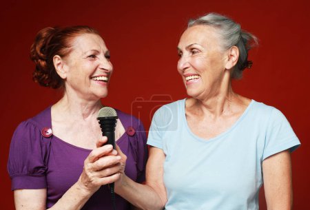 Téléchargez les photos : Deux heureuses amies âgées avec microphone, rient et se préparent pour le karaoké de fête sur fond rouge - en image libre de droit