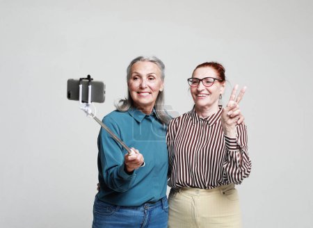 Téléchargez les photos : Deux femmes âgées souriantes prenant un selfie avec un bâton isolé sur fond gris - en image libre de droit