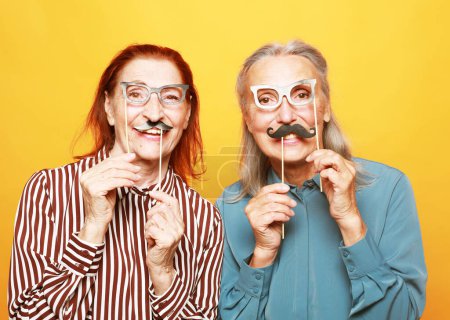 Photo for Funny elderly female friends with fake mustache and glasses, laughs and prepares for party over yellow background - Royalty Free Image