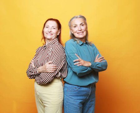 Foto de Dos amigas ancianas sonriendo con los brazos cruzados sobre fondo amarillo. - Imagen libre de derechos