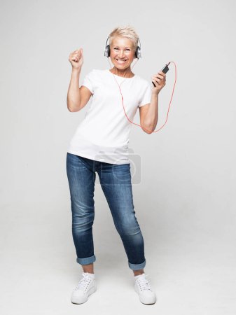 Téléchargez les photos : Portrait de femme âgée aux cheveux courts écoutant de la musique avec écouteurs et smartphone sur fond gris - en image libre de droit