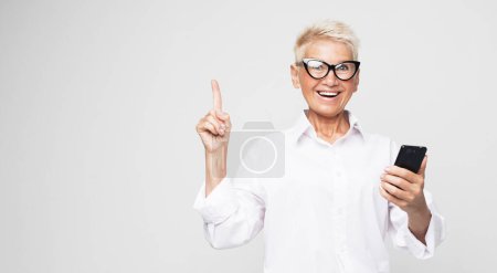 Téléchargez les photos : Belle pensionnée tenant mobile. Une femme âgée moderne tient un smartphone et montre le téléphone, présentant un produit. - en image libre de droit