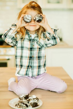 Téléchargez les photos : Style de vie et concept d'enfance drôle de quatre ans fille assise dans la cuisine et manger des biscuits - en image libre de droit
