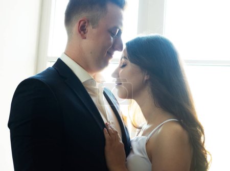 Téléchargez les photos : Jeune couple heureux dans des vêtements de mariage élégant serrant tendrement les uns les autres à la maison. - en image libre de droit