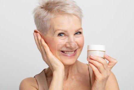 Foto de Sonriente mujer madura de mediana edad de los años 60 poniendo crema facial endurecimiento en la cara mirando a la cámara sobre fondo gris claro. - Imagen libre de derechos