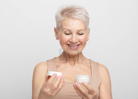 Photo for Smiling 60s middle aged mature woman putting tightening facial cream on face looking at camera over light grey background. - Royalty Free Image