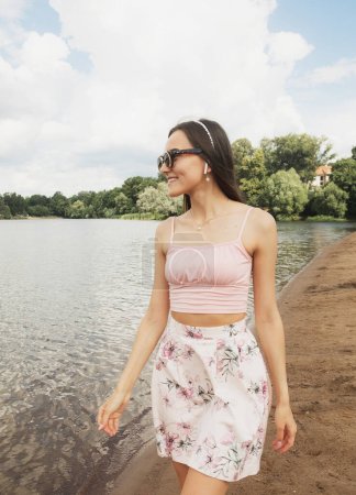 Téléchargez les photos : Belle femme à la mode à l'extérieur. Jeune mannequin femme portant des lunettes de soleil posant près du lac. Style de vie et concept de personnes. - en image libre de droit