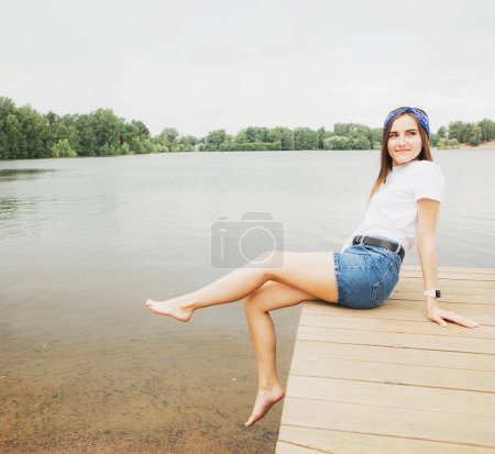 Foto de Una joven alegre en pantalones cortos y una camiseta blanca se sienta en una pasarela de madera cerca del lago - Imagen libre de derechos