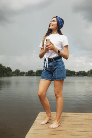 Foto de Una joven alegre en pantalones cortos y una camiseta blanca en una pasarela de madera cerca del lago - Imagen libre de derechos