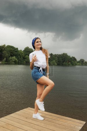 Photo for Young woman listens to music and enjoys vacation, weather before thunderstorm, lake, summer. Lifestyle and people concept. - Royalty Free Image