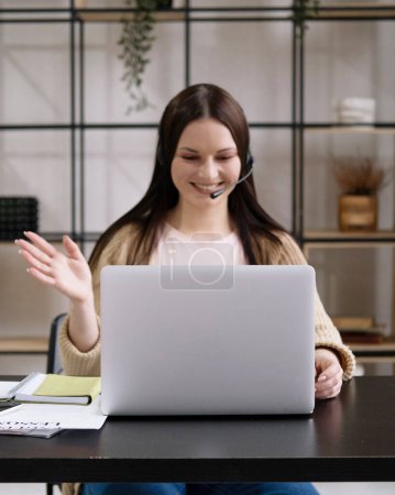 Téléchargez les photos : Jeune heureux heureux gai mignon belle femme d'affaires s'asseoir à l'intérieur dans le bureau en utilisant un ordinateur portable avec casque. - en image libre de droit