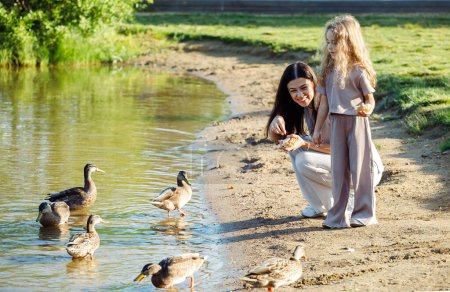 Foto de Una niña alegre de cinco años de edad y su madre están caminando por el lago, mamá y su hija alimentan migas de pan a patos y gaviotas cerca del lago, divirtiéndose en un día de verano. - Imagen libre de derechos