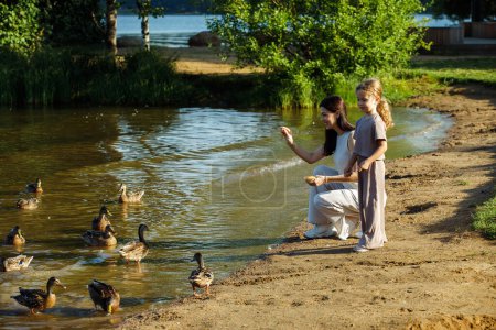 Téléchargez les photos : Une fille gaie de cinq ans et sa mère marchent au bord du lac, maman et sa fille donnent des miettes de pain aux canards et aux goélands près du lac, s'amusant un jour d'été. - en image libre de droit