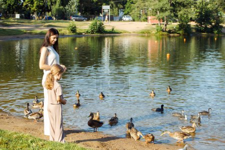 Téléchargez les photos : Une fille gaie de cinq ans et sa mère marchent au bord du lac, maman et sa fille donnent des miettes de pain aux canards et aux goélands près du lac, s'amusant un jour d'été. - en image libre de droit