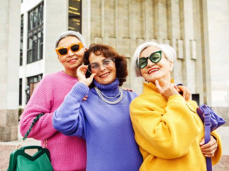 Foto de Tres felices novias jubiladas alegres en suéteres brillantes y gafas de sol caminan juntas en la ciudad en un día de otoño. - Imagen libre de derechos