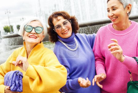 Photo for Three happy cheerful pensioner girlfriends in bright sweaters and sunglasses walk together in the city on an autumn day. - Royalty Free Image
