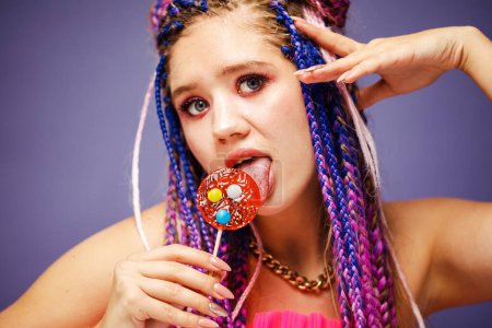 Photo for Young smiling woman with dreadlocks hairstyle and creative make-up in doll style with candy over purple background - Royalty Free Image