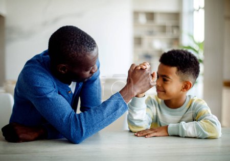 Photo for Happy african american father competing in arm-wrestling with his son, enjoying time together at home. Lifestyle concept. - Royalty Free Image