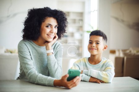 Téléchargez les photos : Portrait de jeune mère afro-américaine tenant smartphone lecture enseignement petit garçon dans le salon. École à domicile apprendre de l'enseignant, l'éducation famille heureuse. - en image libre de droit